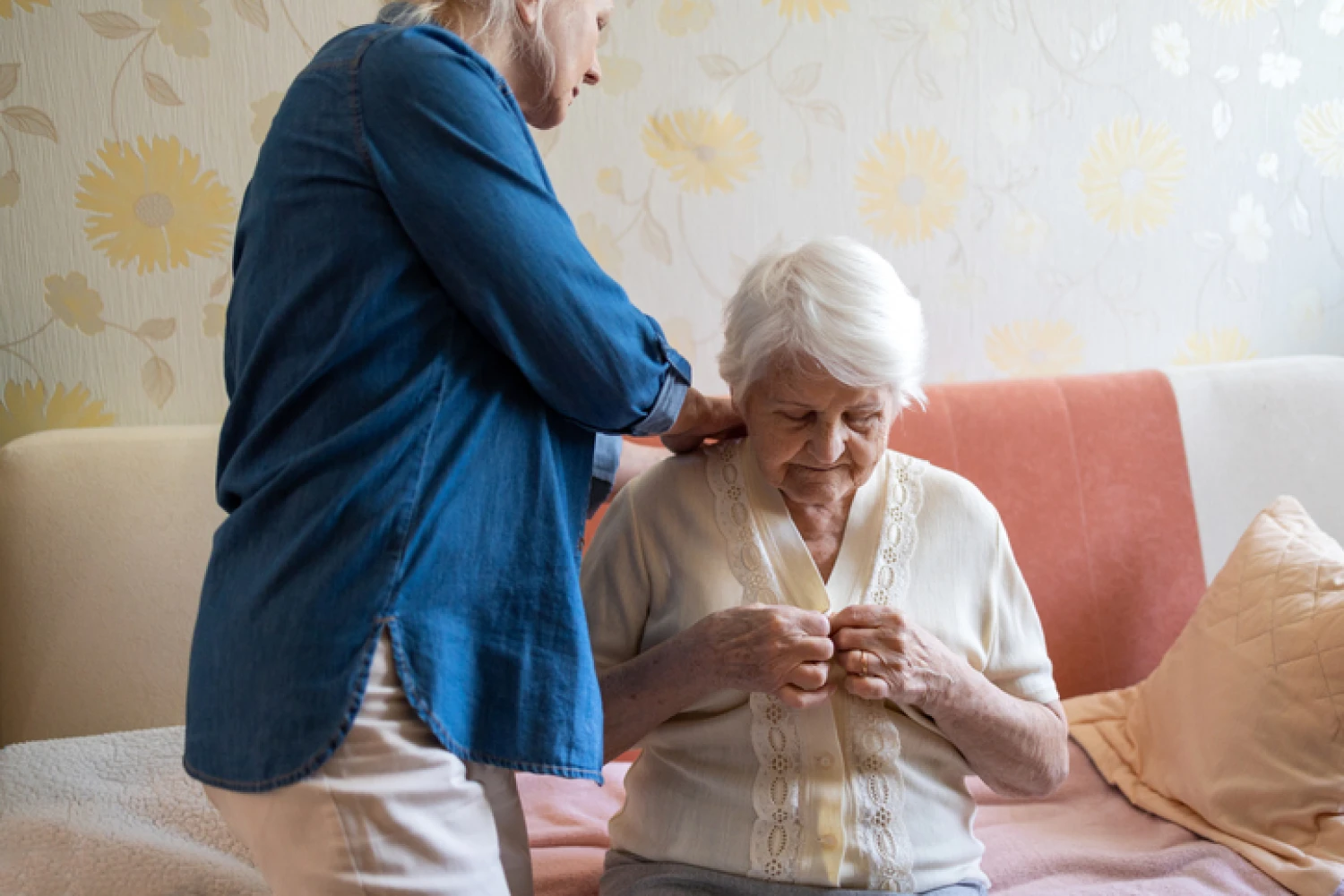 A caregiver helps an elderly woman button her shirt, demonstrating support with dressing as a part of personal care.