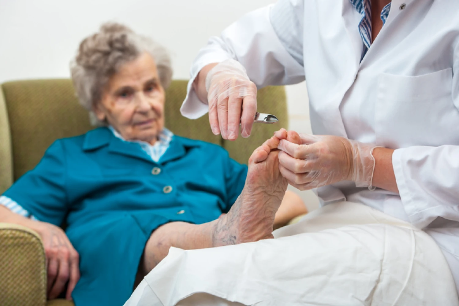A caregiver in gloves provides foot care to an elderly woman sitting in an armchair, focusing on maintaining hygiene and well-being.
