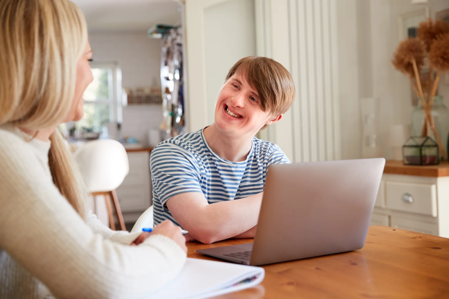 Boy sitting behind laptop smiles at blonde lady with a notebook.