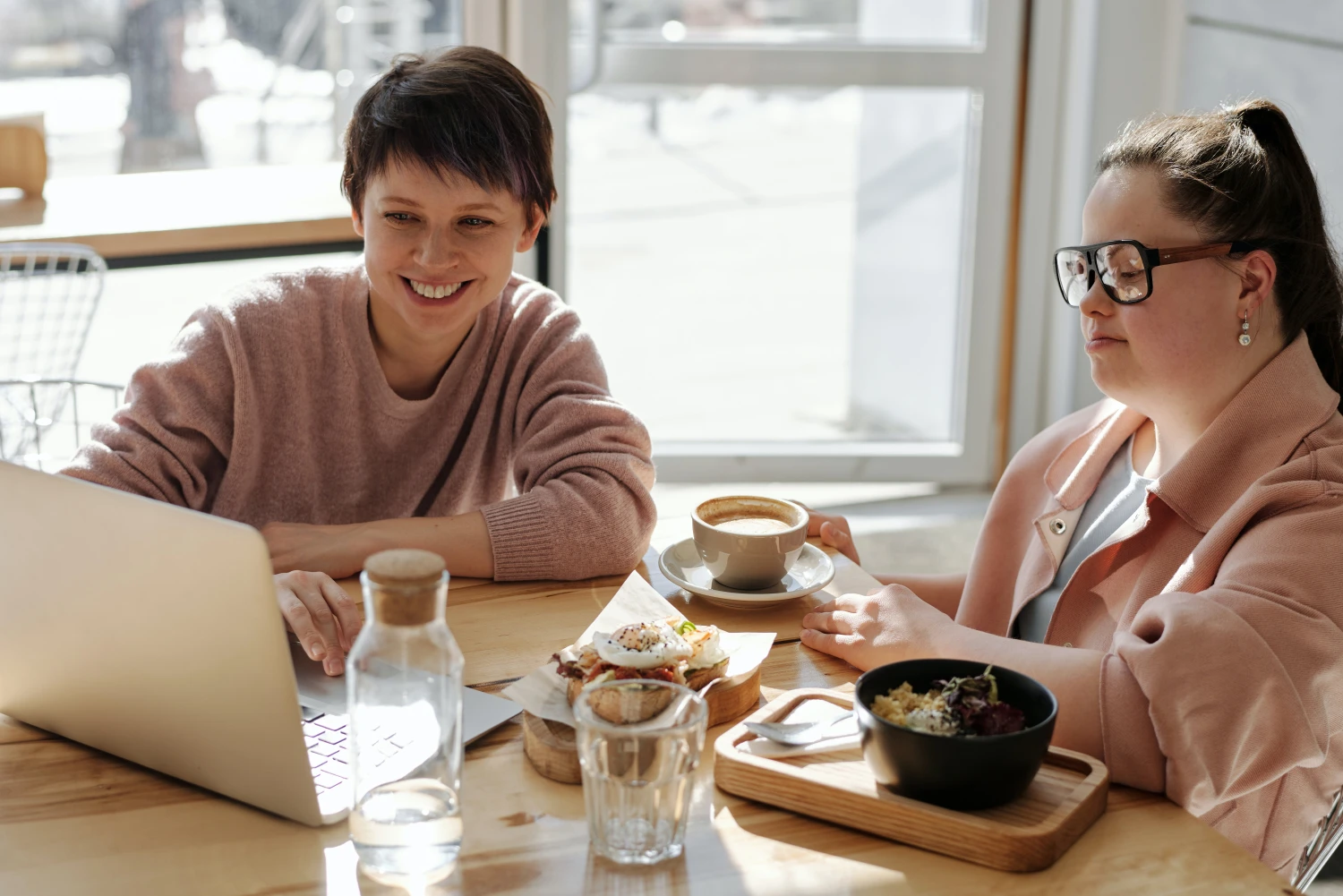 A lady with Down Syndrome sitting at a coffee table with a laptop and a friend - 5 Tips to Know Before Your NDIS Plan Review - Focus Care